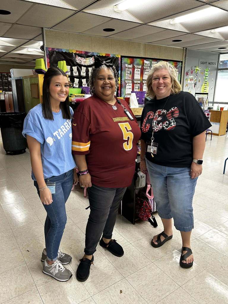 Three women stand indoors smiling at the camera. The woman on the left wears a light blue T-shirt and jeans, the middle woman wears a maroon sports jersey with "COMMANDERS" and the number 5, and the woman on the right wears a black T-shirt with a red and white design and rolled-up jeans. The background shows a colorful bulletin board and classroom or office supplies.