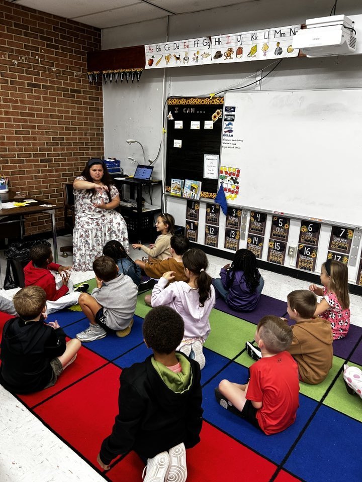 teacher sits at front of room teaching while students look on while sitting on the carpet