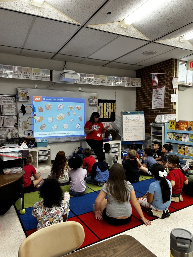 A teacher stands at the front of the room next to an active board with students looking on as they sit on the carpet.