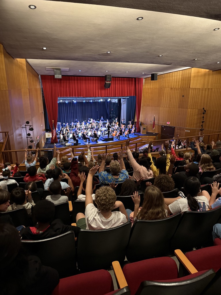 A large auditorium filled with people watching a live orchestra performance on stage. The stage is framed with red curtains, and musicians with string and wind instruments are seated under stage lights. The audience occupies most of the seats, and the photo is taken from an upper balcony view, showing both the performers and the crowd.