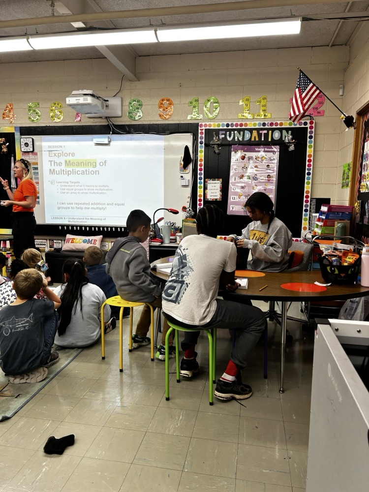 student intern helps a group of students as the teacher instructs students at the front of the room
