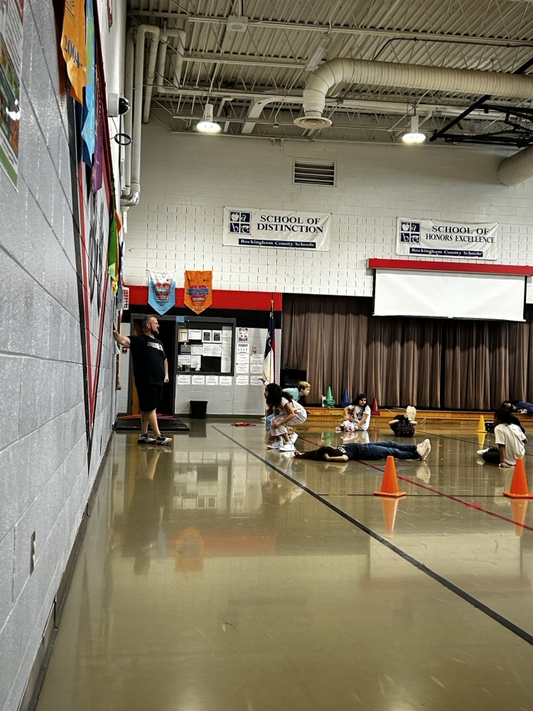 the gym teacher explains the exercises to the group of students sitting on the floor watching
