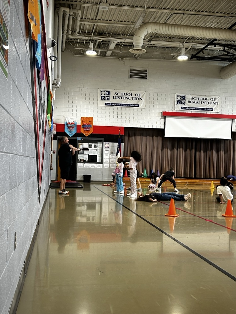 the gym teacher explains the exercises to the group of students sitting on the floor watching