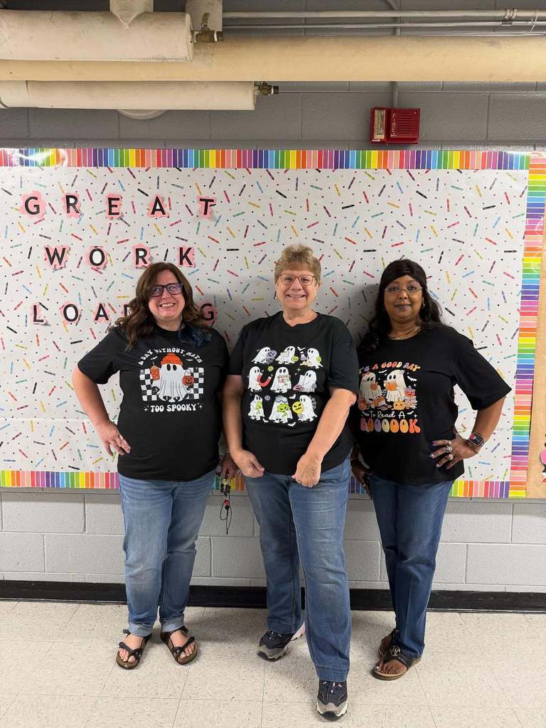 Three ladies stand in front of a bulletin board wearing their black Halloween shirts with ghosts on them.
