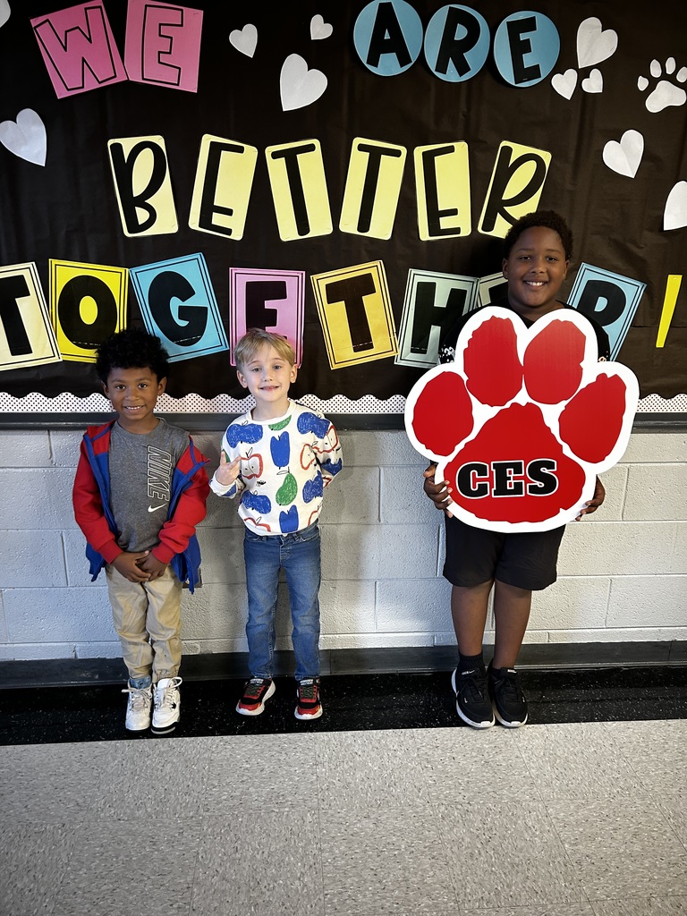 Three boys stand in front of a bulletin board. One stands holding a red paw print with the letters CES on it.