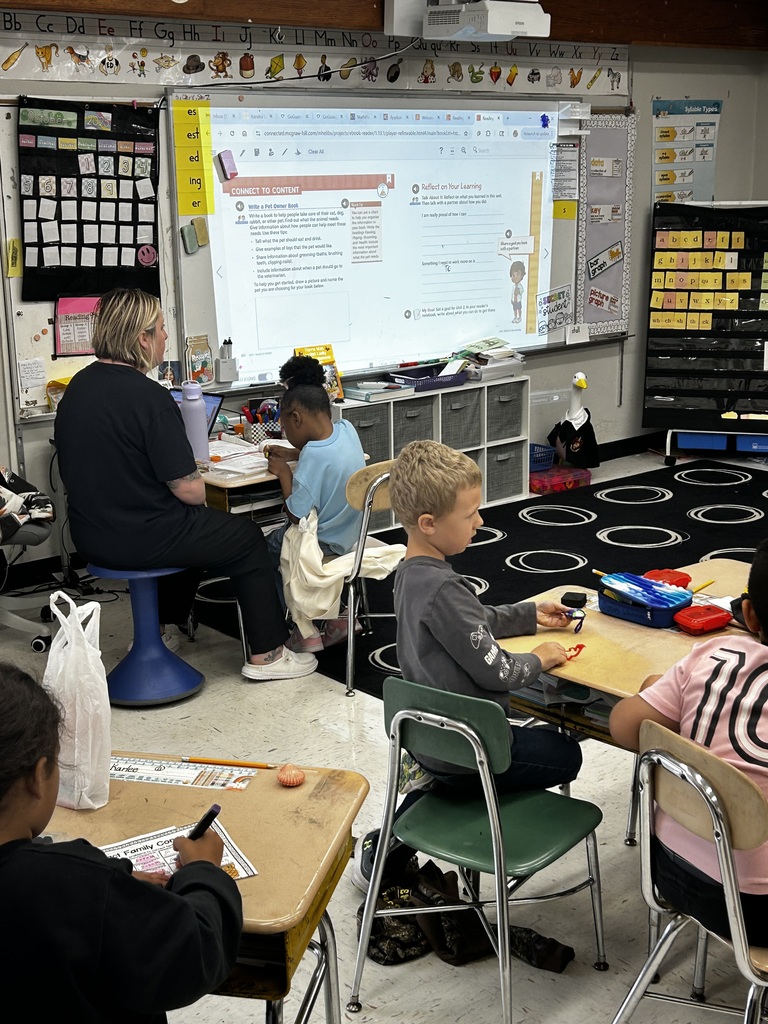 teacher sits at front of room working with another student at the board