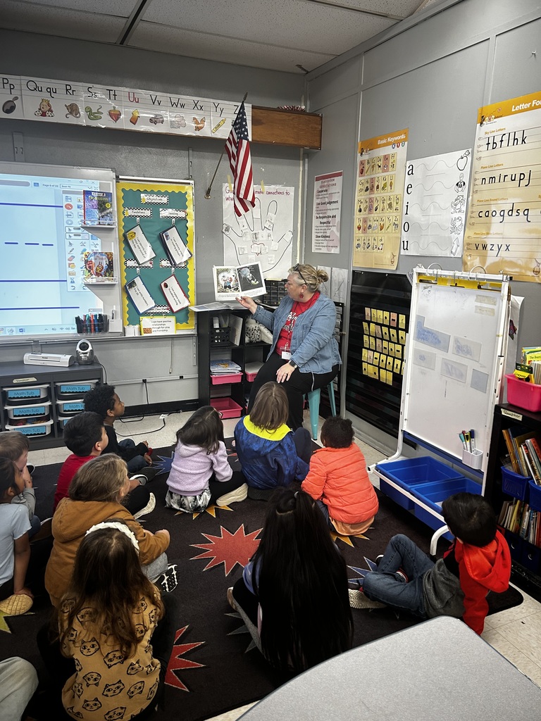 Teacher sits at front of the room reading book to students as students sit on the carpet and look on