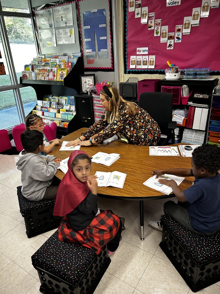 teacher sits at kidney table working with students as they read paper books