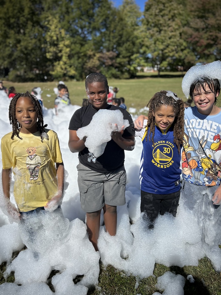 four male students stand in foam. One holds a huge amount of foam and another has foam on his hand. 