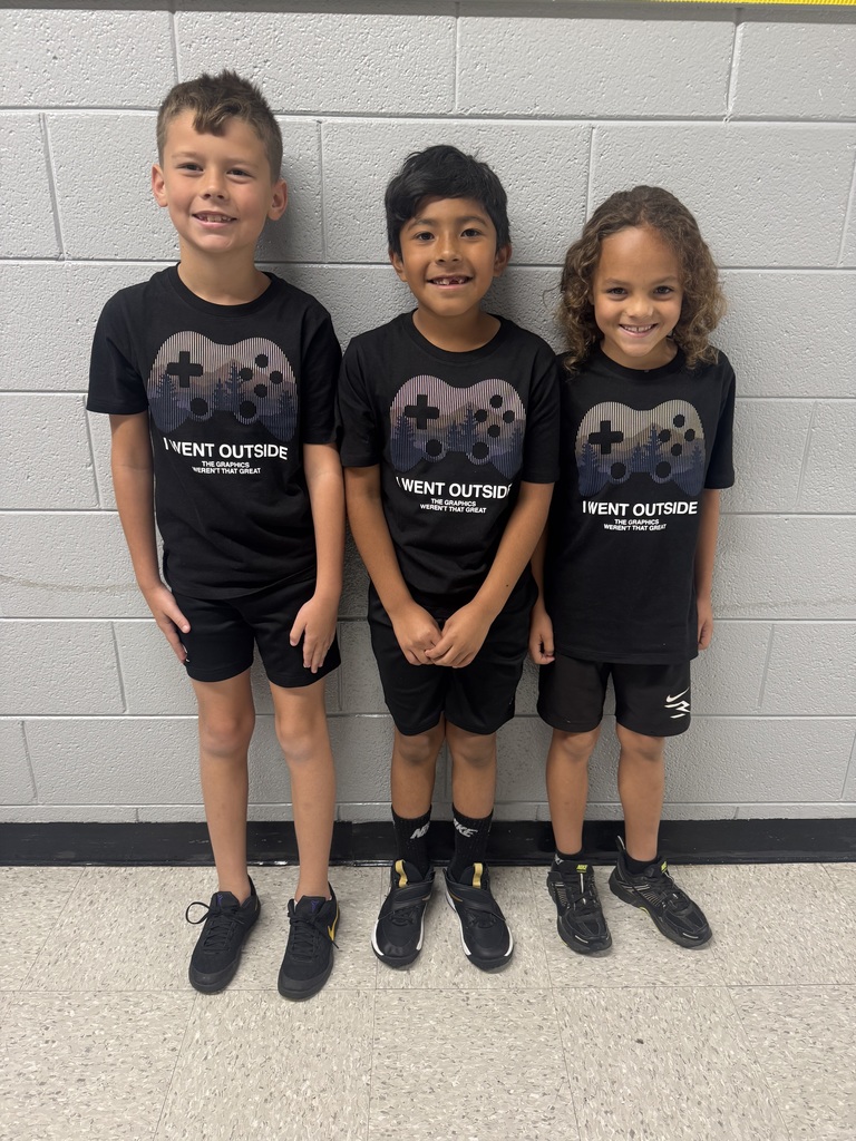 three boys stand together wearing matching black shirts with a video game controller on it, black shorts, and black socks and shoes. 