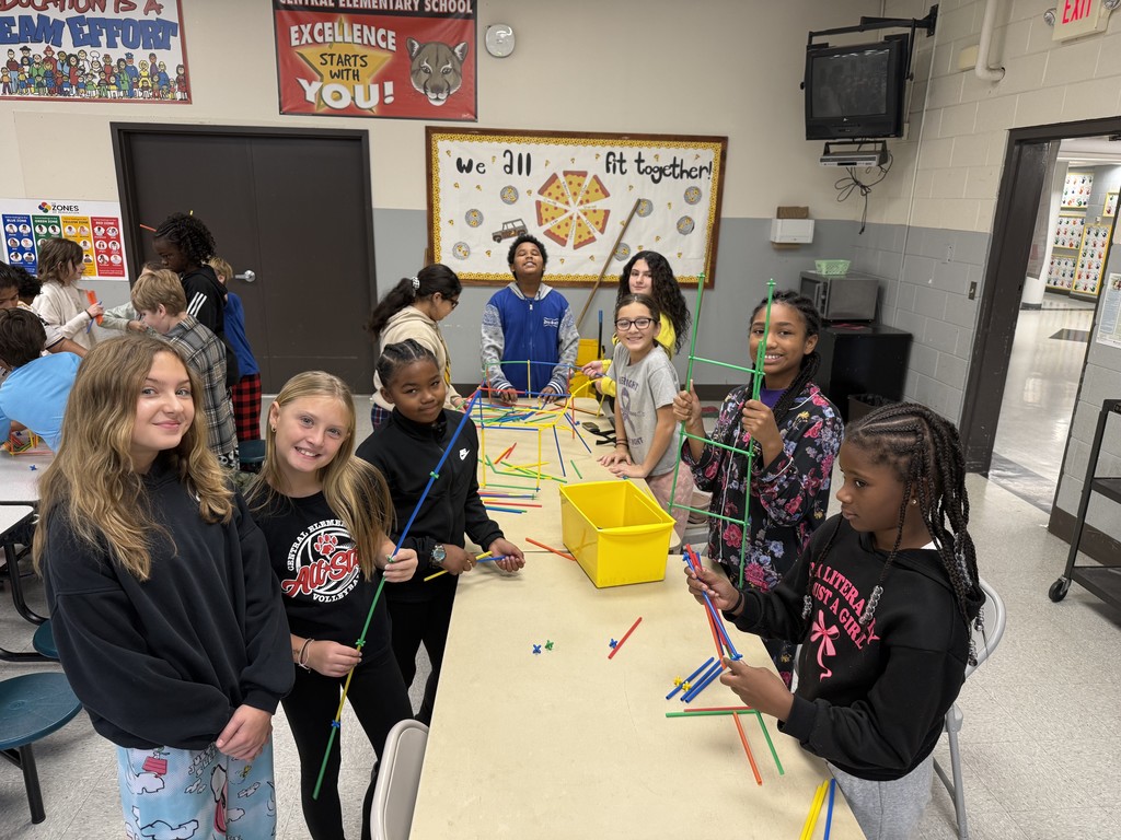 Group of students stand at table building straw structures. 