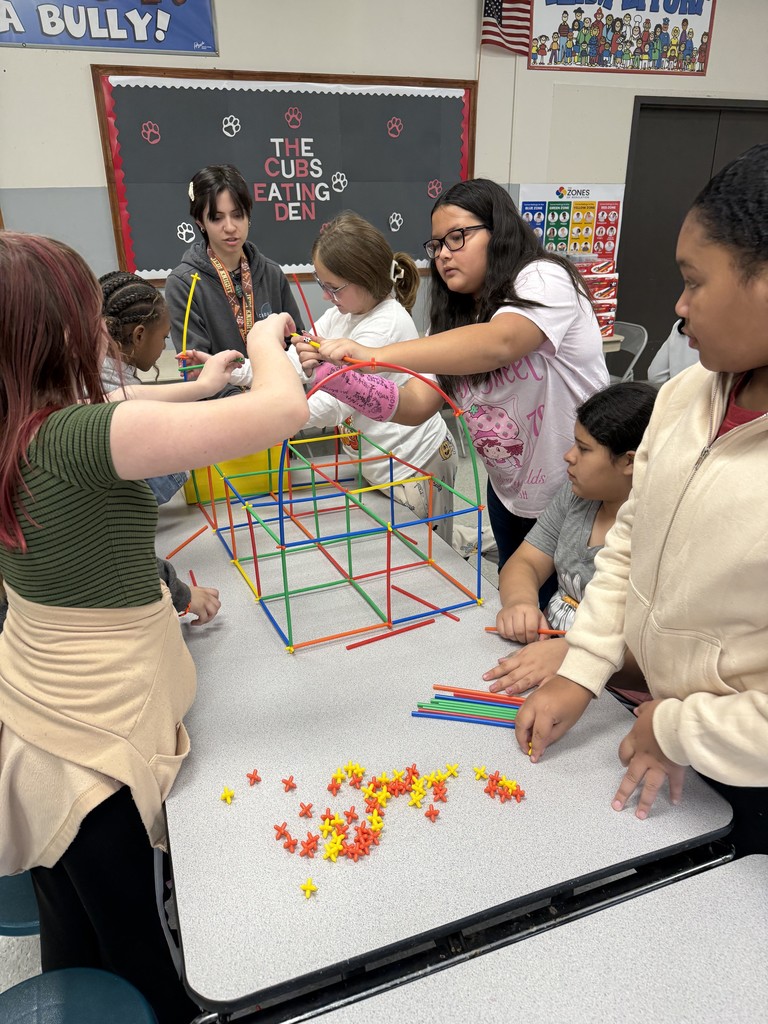 Group of students stand around table building straw structure. 