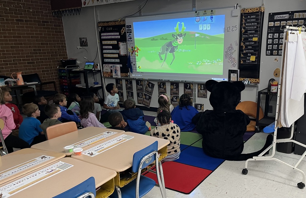 Students sit on carpet watching program on active board while black panther cub sits on carpet too. 