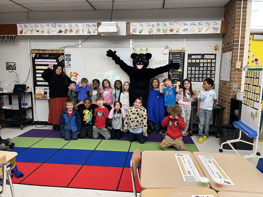Students and teacher stand at the front of the room with black panther cub mascot. Mascot is seen with hands in the air.