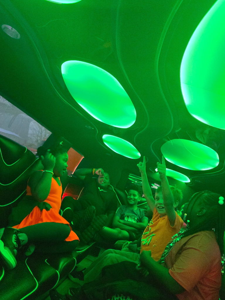 Students sit in group with hands raised inside of limo with green lights illuminating background