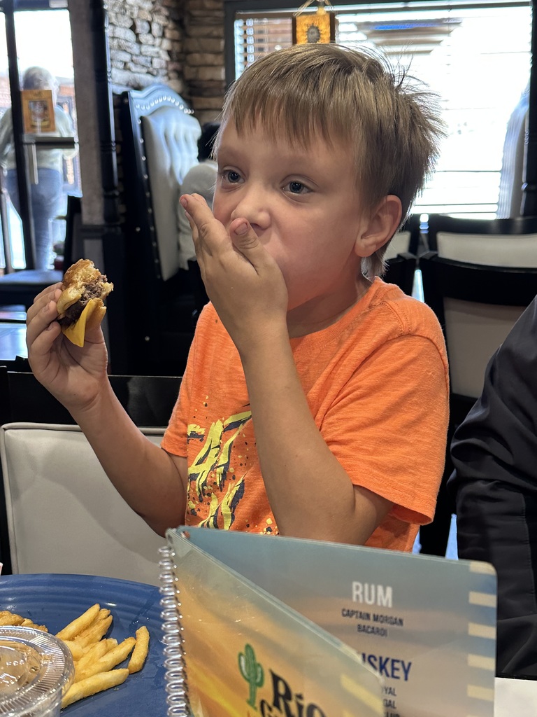 male student in orange shirt eats cheeseburger and covers mouth with his hand. 