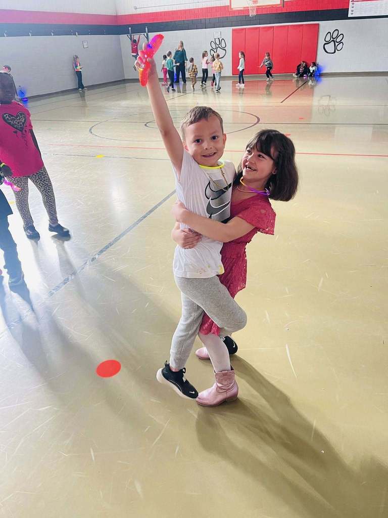 a girl holds a boy up in the air. he has his arm raised with neon sunglasses in his hand. 