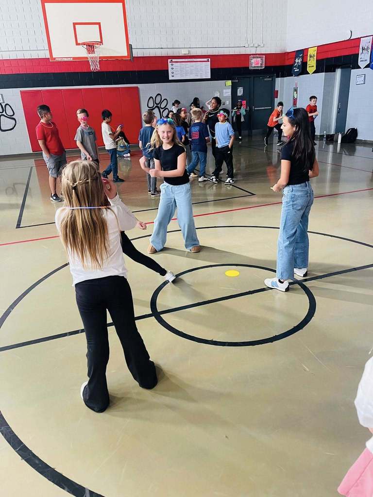 three girls stand in circle in the gym. one dances while the others watch. 