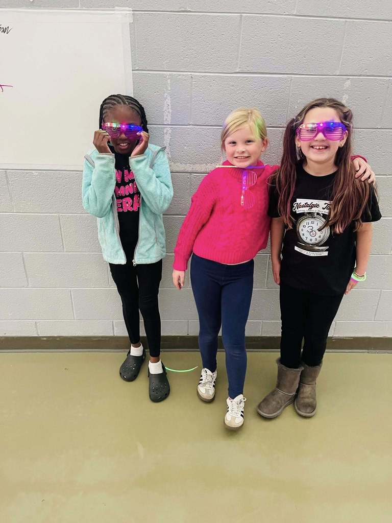 three girls stand together smiling. two wear glow in the dark sunglasses. 
