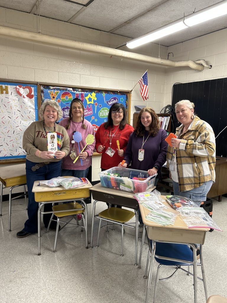 “A group of five adults stand smiling in a classroom behind a set of student desks. They are holding colorful educational craft materials, and there are bins and bags of supplies on the desks in front of them. Behind them is a bulletin board decorated with bright colors and an American flag on the wall.”