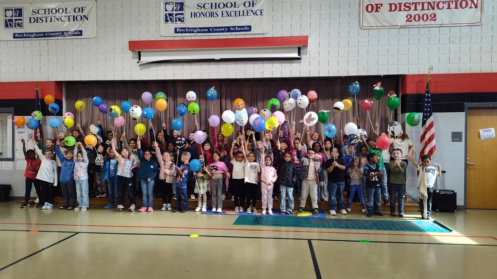 A large group of elementary-aged children stand together on a gymnasium stage, each holding a colorful balloon decorated with different drawings and designs. The kids are smiling and raising their balloons high in the air. Behind them hangs a curtain, and above are school banners recognizing distinctions and honors. An American flag stands to the right side of the stage. The gym floor and walls are visible in the foreground.