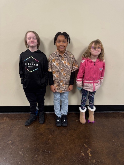 “Three young children stand side by side against a beige wall on a polished brown floor. The child on the left wears a pink jacket, patterned leggings, and tan boots with white fur trim. The child in the middle wears a camouflage-print shirt over a black long-sleeve top, light jeans, and black shoes. The child on the right wears a black hoodie with a geometric design, black pants, and black shoes. All three children are smiling and facing the camera.”