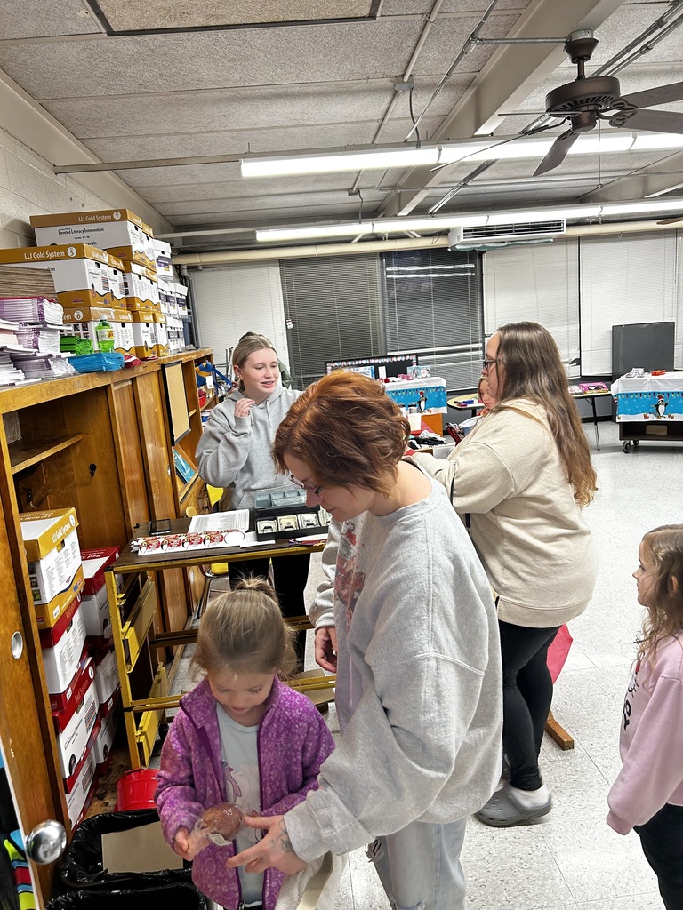 Three adults stand in a classroom. One is helping a female student purchase items in the holiday shoppe. The other two are standing and talking to each other. A female student stands behind them looking on. 