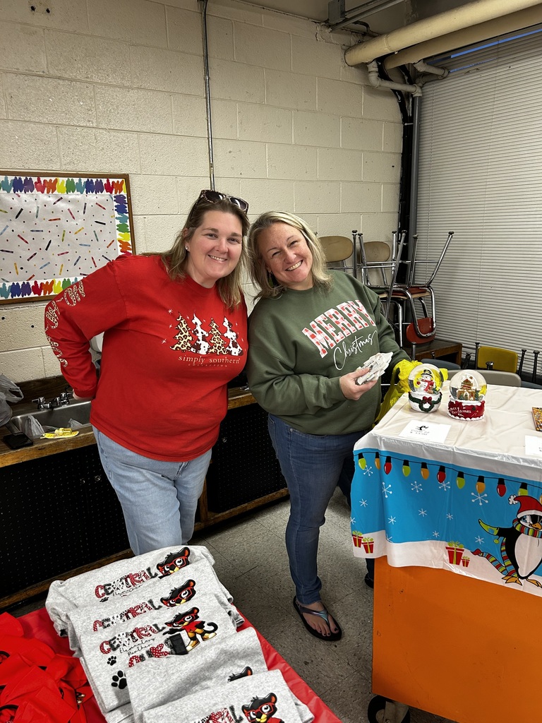 Two women stand, leaning together, in the holiday shoppe. There is a table in front of each filled with merchandise for students to purchase. 