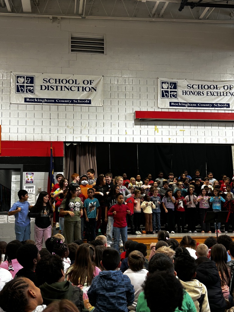 Here is alt text for the image:  **Alt text:** A large group of elementary students stand on risers in a school gymnasium, performing together while an audience of students sits on the floor watching. Several students in the front row of the performers have stepped forward and are doing hand motions as part of the performance. Above the stage, two banners hang on the brick wall reading “School of Distinction” and “School of Honors Excellence.” The gym is brightly lit with exposed ceiling beams.