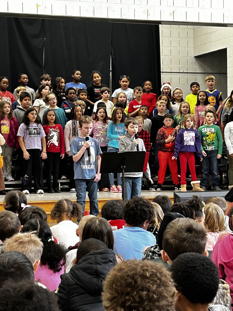 A large group of third-grade students stand on risers on a school stage, singing during a performance. Two boys stand at microphones in front of the group, reading or speaking from a music stand. In the foreground, many younger students sit on the floor watching the performance. The background includes black stage curtains and a light-colored brick wall.