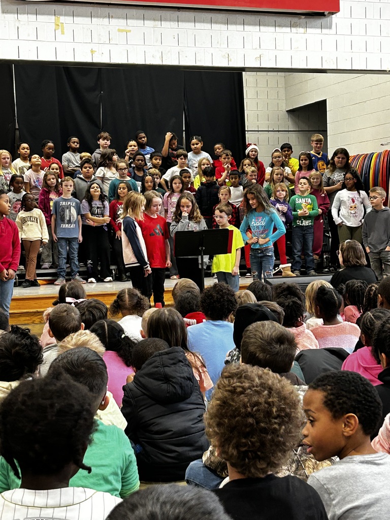 A large group of third-grade students stand on risers on a school stage, singing during a performance. Four students stand at microphones in front of the group, reading or speaking from a music stand. In the foreground, many younger students sit on the floor watching the performance. The background includes black stage curtains and a light-colored brick wall.