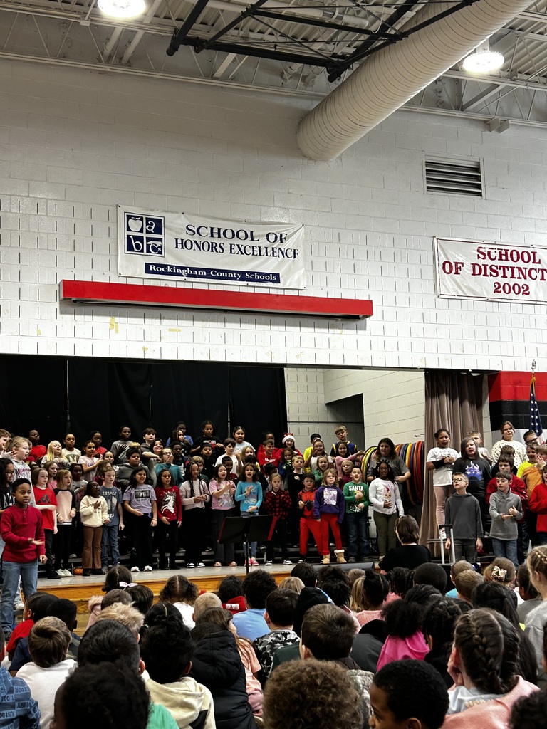 A large group of third-grade students stand on risers on a school stage, singing during a performance.  In the foreground, many younger students sit on the floor watching the performance. The background includes black stage curtains and a light-colored brick wall.