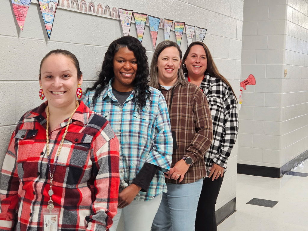 Four women stand side‑by‑side in a school hallway, smiling at the camera. From left to right: a woman with light hair pulled back wearing a red/black/white plaid shirt, wooden‑bead necklace and red/yellow patterned earrings (ID badge visible); a woman with long dark wavy hair wearing a blue/white plaid shirt over a dark top; a woman with lighter hair wearing a brown/white plaid shirt and a watch; and a woman with long brown hair wearing a black/white plaid shirt over dark pants. They stand in front of a white brick wall decorated with a colorful triangular‑flag bunting and student artwork flags that repeat the phrase “ALL ABOUT ME” (the leftmost flag reads “MY FAVORITE…” with a drawing). 