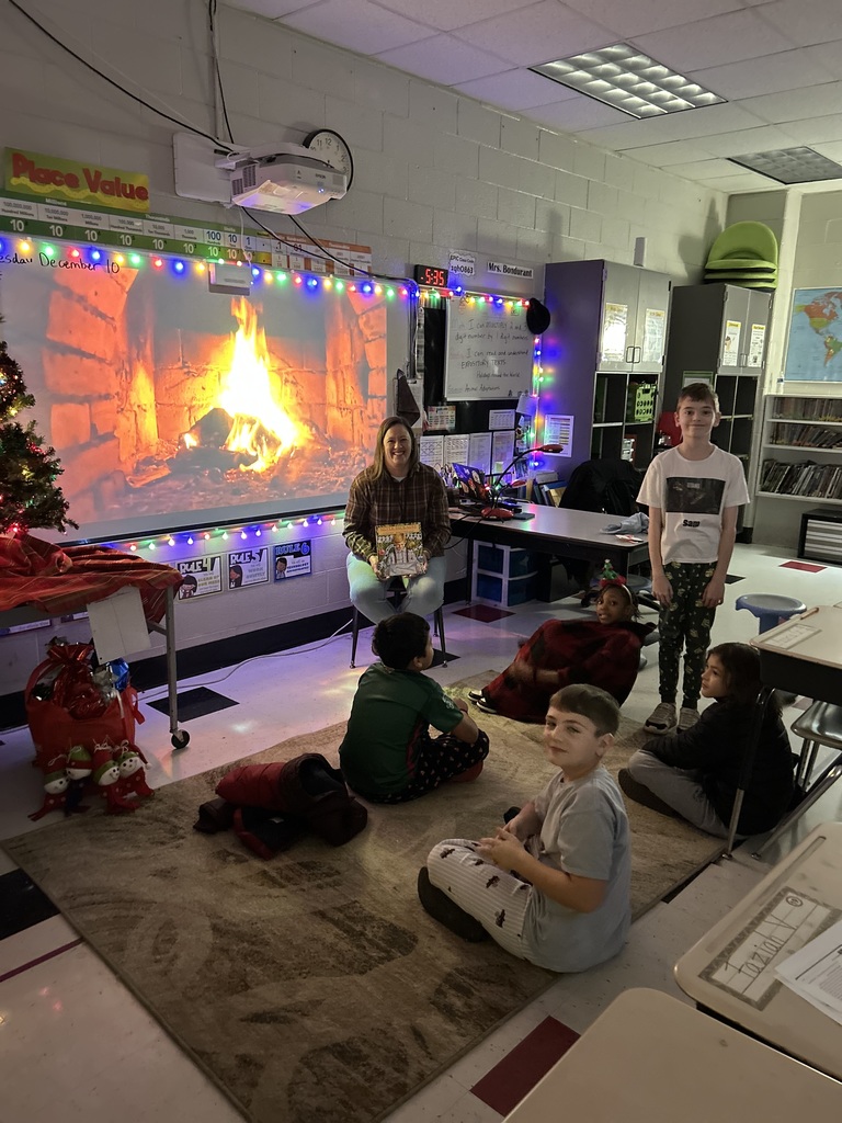 A classroom decorated with colorful holiday string lights and a large projected fireplace scene. A teacher sits on a chair at the front of the room holding a picture book, smiling as several young students sit on a rug facing her. Some students wear festive pajamas and one is wrapped in a blanket. The room is dimly lit except for the fireplace projection and string lights, creating a cozy, warm atmosphere. Classroom furniture, posters, and cabinets are visible in the background.