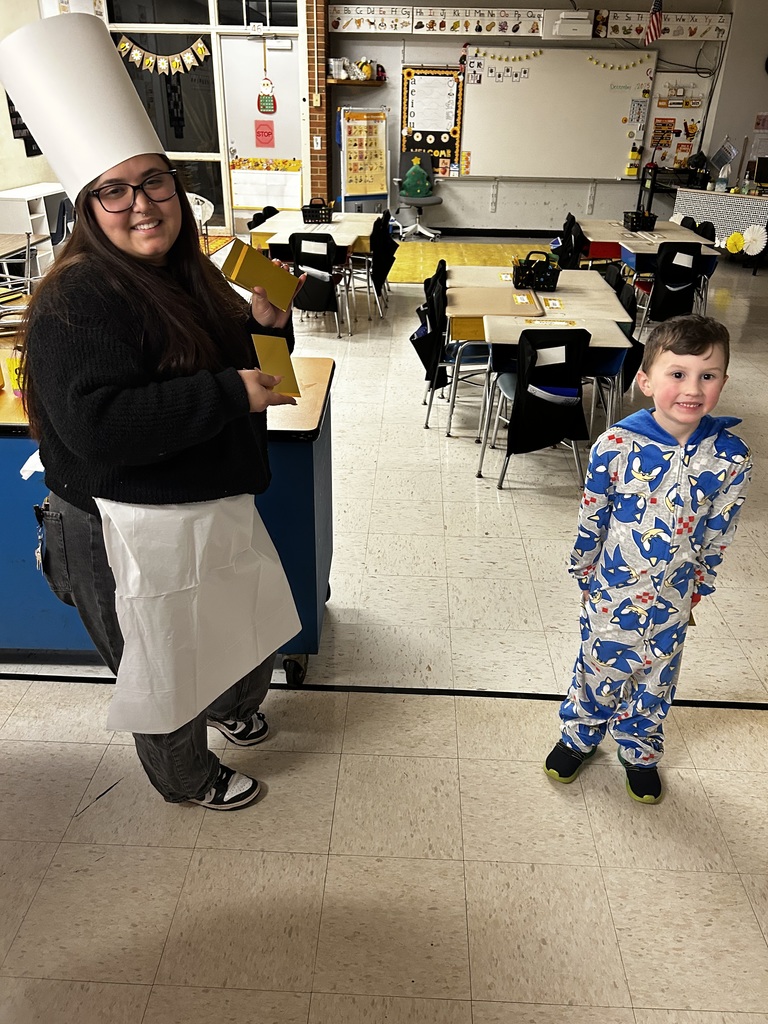 A classroom portrait of an adult woman and a young boy standing side by side. The woman on the left wears a tall white chef’s hat, glasses, a black long-sleeve sweater, dark gray pants, and a white apron; she smiles and holds two small yellow rectangular boxes. The boy on the right wears a blue-and-white Sonic the Hedgehog patterned full-body pajama suit and smiles at the camera. Behind them are light-colored student desks with black chairs on a beige tiled floor. 
