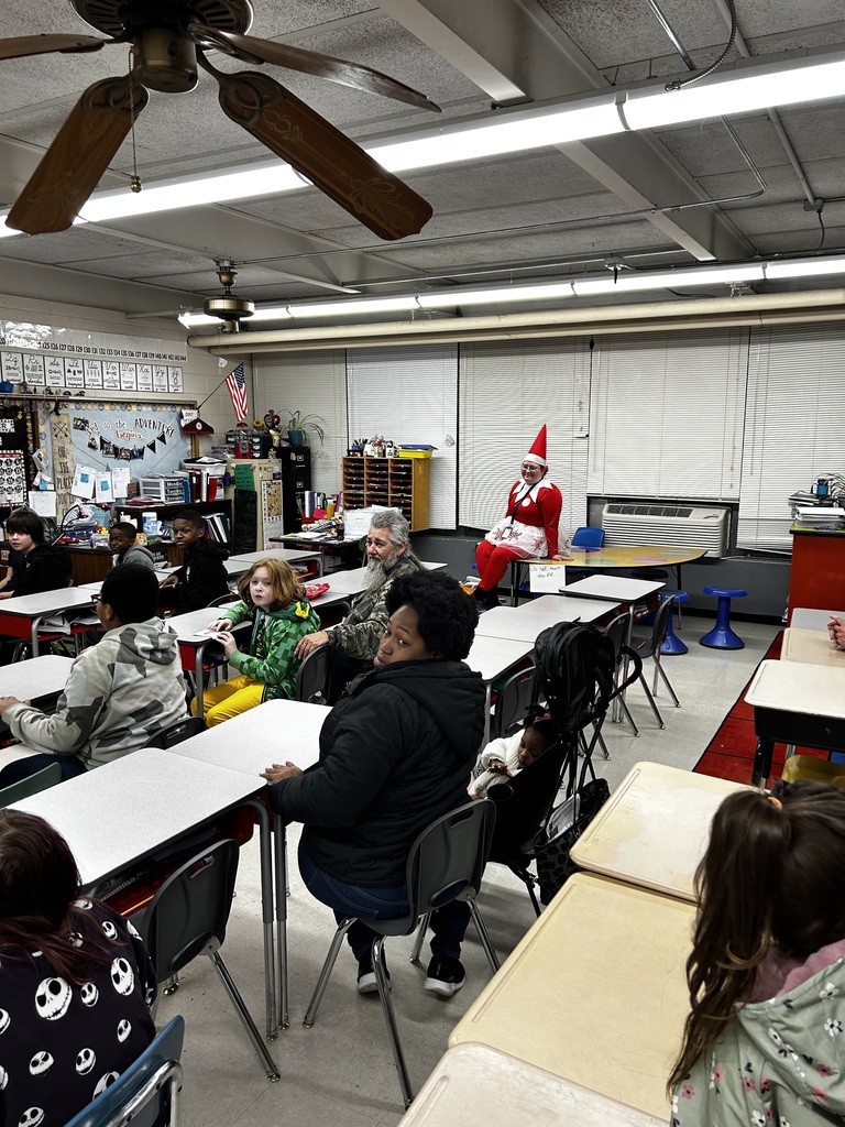 Classroom scene with students and adults seated at individual desks facing the front. Overhead are fluorescent lights and two wooden ceiling fans. In the foreground a young person in a dark jacket and glasses stands facing the room; nearby a student with reddish-brown hair wears a purple long-sleeve shirt patterned with small white Jack Skellington–style ghosts and sits at a front desk facing away from the camera. Adults sit among the students — one with long gray hair and a beard at center-right, another with dark hair seated beside a baby in a black car seat or stroller. On the far right an individual dressed as a festive elf in a red suit with white trim and a pointy hat is seated on a shelf or counter. Light-colored walls surround the room; the left wall features a large whiteboard with a projector above it and a long strip showing a number line and alphabet. To the right of the whiteboard are decorated bulletin boards, shelves, and an American flag. The floor is light tile.