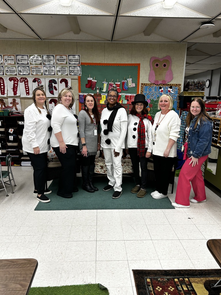 Seven women pose together in a brightly decorated elementary classroom, dressed in coordinated holiday-themed outfits that resemble snowmen (white tops with large black “buttons,” scarves, pom‑pom headbands, and a black top hat on one wearer). From left to right they wear combinations of white tops, black pants, a gray knit dress, a cream sweater, and a blue denim jacket with bright pink pants; several wear headbands or festive accessories and one woman has an identification badge. They stand on a dark green rug in front of classroom displays and furniture; behind them are shelves, a patterned bench, and bulletin boards decorated with stockings and a pink owl cutout.