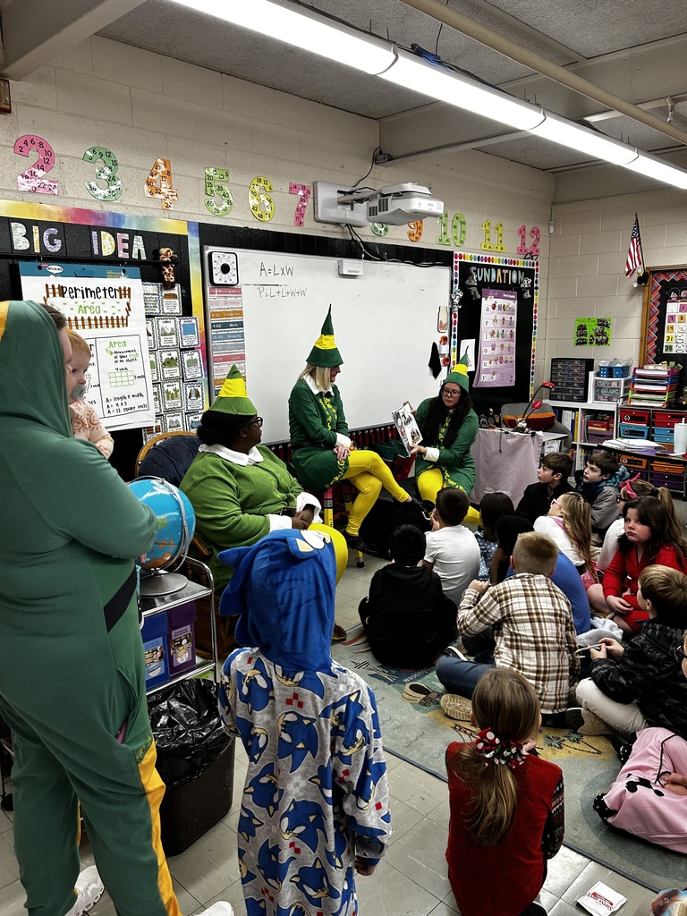Bright elementary classroom during a group reading or storytelling session. In the foreground about 15–20 young children sit on a colorful rug, facing several adults dressed as elves; two elf-costumed adults sit at the front (one on the right is holding an open book and appears to be reading) while a third elf stands at left holding a baby. One child stands at left wearing a blue Sonic the Hedgehog onesie. Overhead fluorescent lights and a ceiling-mounted projector illuminate the room.