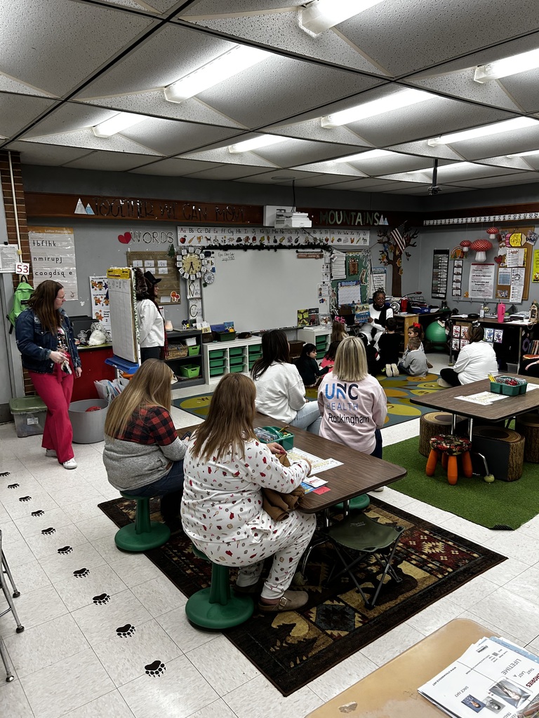 Interior classroom scene with adults and young children gathered for a group activity. In the foreground several adults sit at small dark-wood tables with green stools or on the floor; one adult wears patterned pajamas, another a red-and-black plaid shirt, and a third a pink shirt reading “UNC HEALTH Rockingham.” Children and more adults sit on rugs and low wooden-stump seats in the midground facing a woman leading the activity. The room has light gray walls, a tiled floor with visible bear-paw footprints, fluorescent ceiling lights, and a large whiteboard at the back framed by an alphabet chart (“Aa Bb Cc … Zz”).