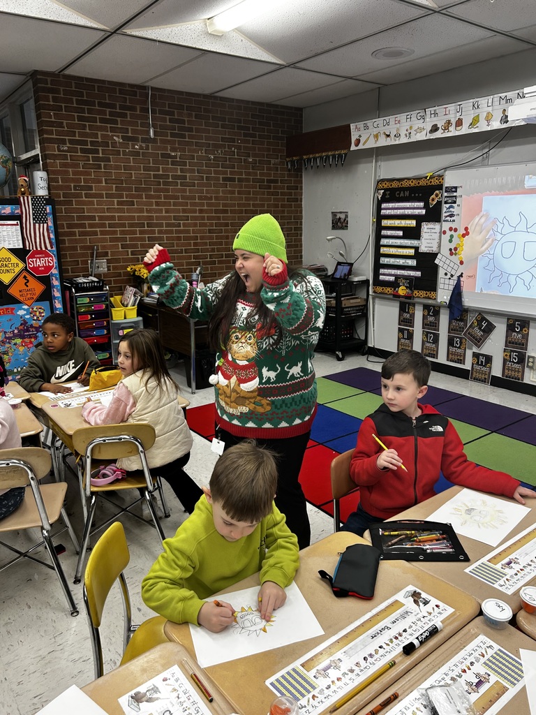 A teacher wearing a holiday sweater raises both arms excitedly while students continue working at their desks coloring pictures. The classroom features a colorful rug, desks with pencils and crayons, and learning displays on the walls.