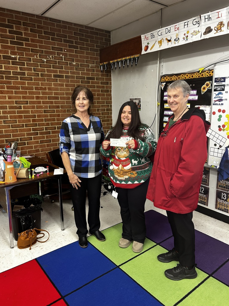 Three adults stand together in an elementary classroom smiling for a photo. The person in the center holds a paper or certificate. Behind them are classroom supplies, a brick wall, and educational posters.