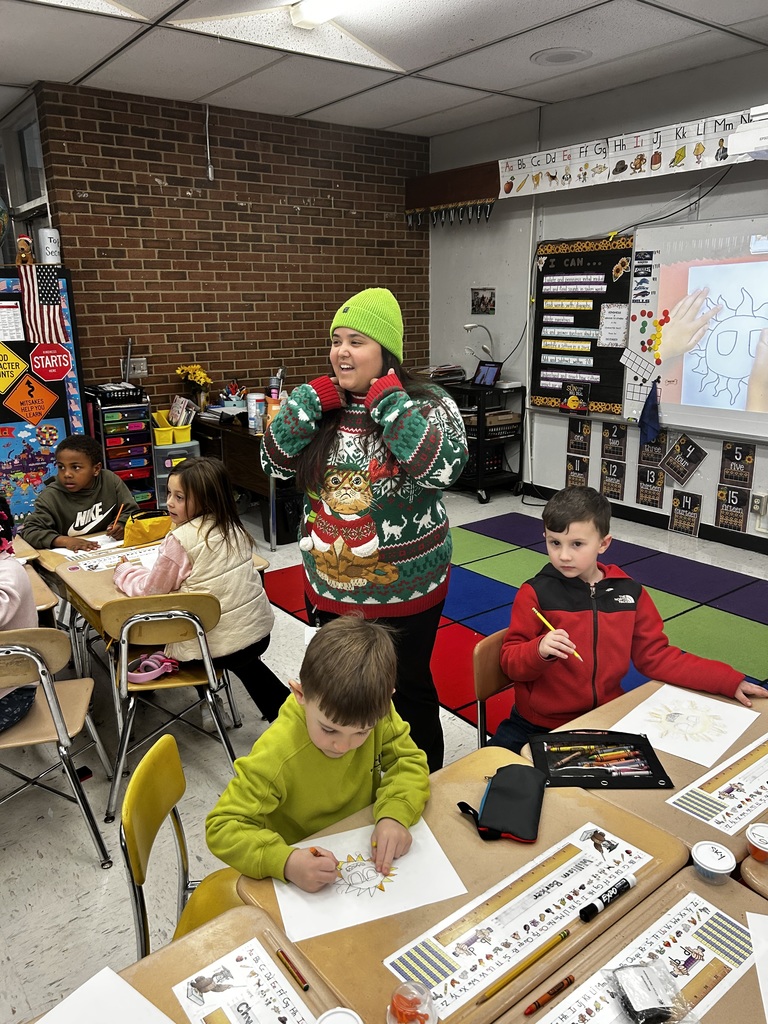 An elementary classroom where several students sit at desks coloring drawings. A teacher wearing a festive holiday sweater and a bright green beanie stands smiling near the students. The classroom includes colorful rugs, posters, and learning materials on the walls.