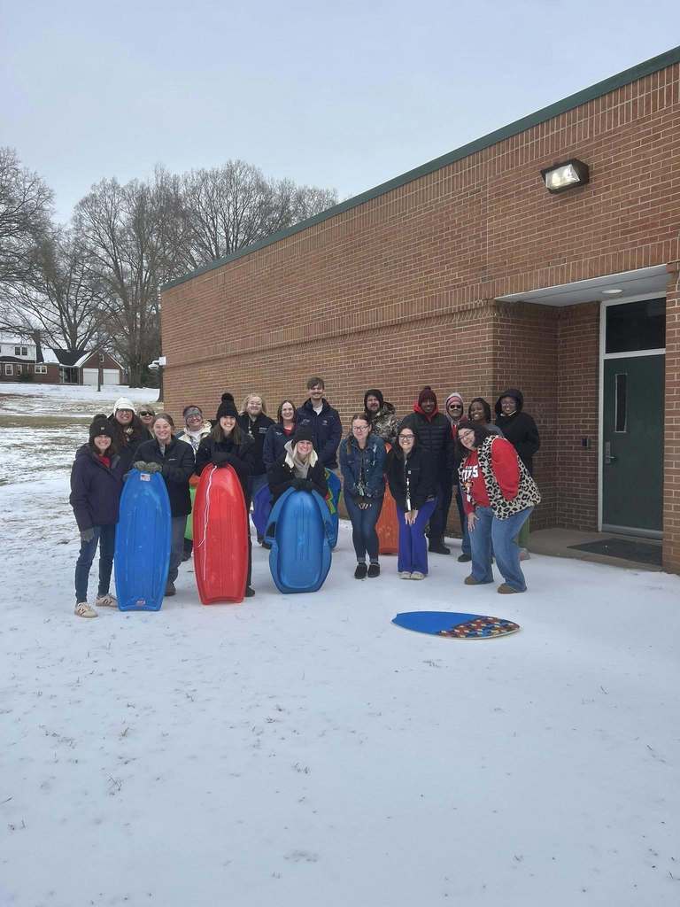 A large group of adults stand smiling outside a brick school building on a snowy day, bundled in winter coats and hats. Several people hold colorful plastic sleds—red, blue, and green—while snow-covered grass and bare trees are visible in the background.
