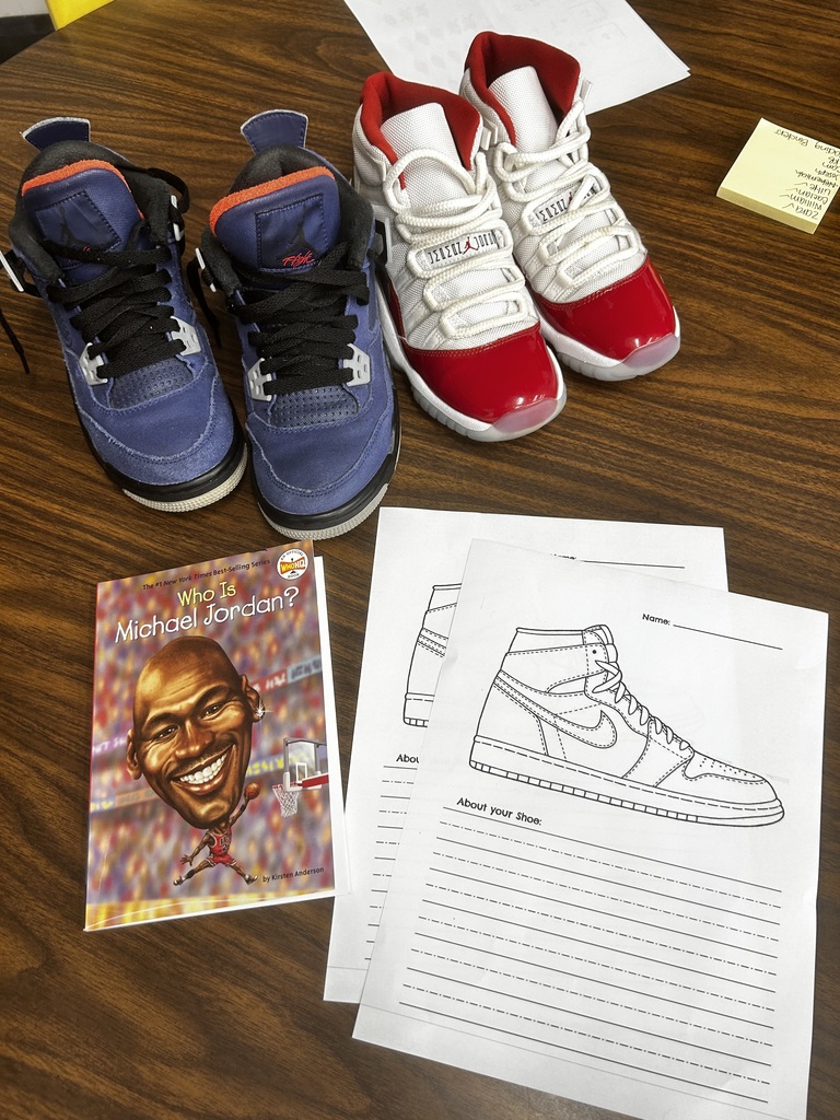 Flat lay of student materials and shoes: two pairs of children’s high-top sneakers (navy/black with orange accents on left; white/red patent on right) arranged on a wooden surface. Below the left shoes is a book titled “Who Is Michael Jordan?” Two identical worksheets with an outline of a high-top sneaker and blank writing lines sit to the right. A yellow sticky note with a short list of names is in the top right corner.