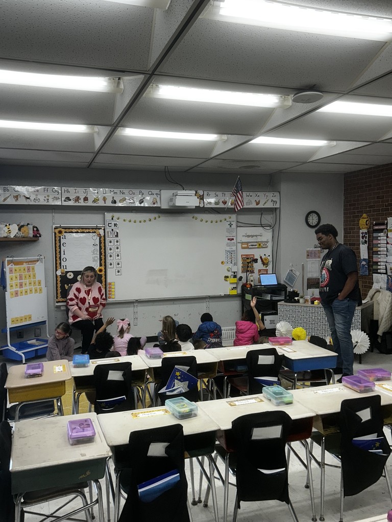 Whole-group instruction on the floor: a female adult in a white sweater with red hearts sits on the floor leading children who raise hands; a male adult stands nearby. The room shows an alphabet border, a vowel chart, a whiteboard, a “FEBRUARY 2024” chart, an American flag, and a ceiling-mounted projector. Desks with colored supply containers and student storage pockets appear in the foreground.