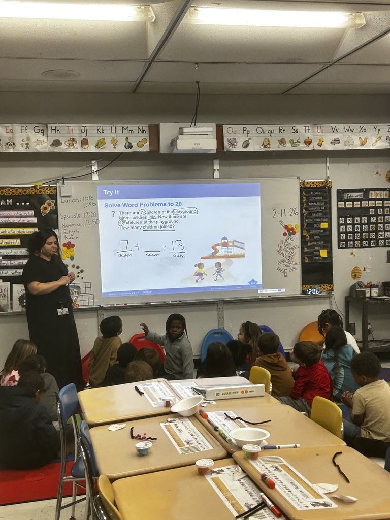 A classroom during a math lesson: a teacher in a black top and skirt stands left of a large projected screen showing a word problem—“There are 7 children at the playground. More children join. Now there are 13 children. How many joined?” with the equation “7 + ____ = 13.” Students sit on the floor facing the screen; desks with markers, containers, and worksheets fill the foreground. Walls display an alphabet chart, a February calendar, a class schedule, and posters. A whiteboard lists “Lunch: 11:15–11:45” and “Specials: 12:20–” with names. Date and time on the board read “2-11-26” and “12:06.” A bulletin board with phonics notes is partially visible.