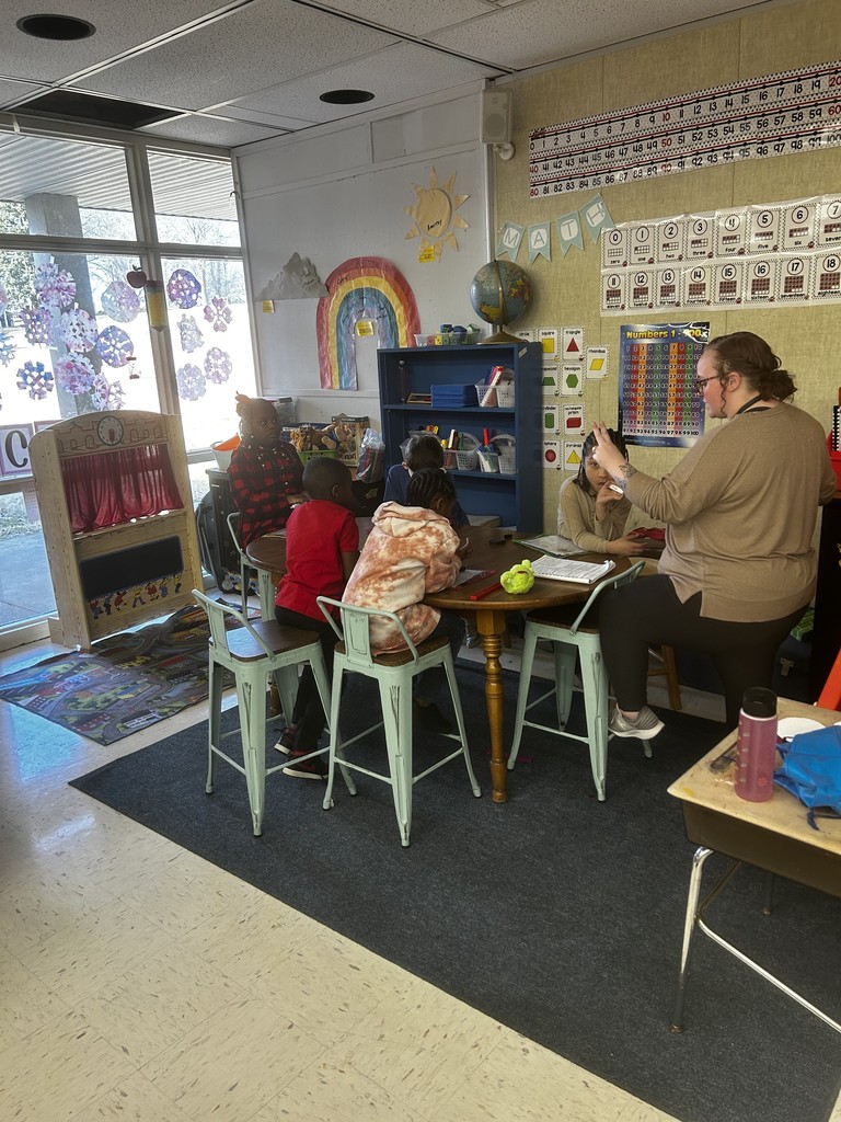 Math center at a round table: a teacher and several children sit at a round table working together. Walls are decorated with a number line (0–100), a “MATH” banner, number-word cards (one–eighteen), a shapes poster, and a 1–100 chart. A hand-drawn sun, puppet theater, bookshelf with a globe, and classroom bins are visible; windows show paper snowflake decorations.