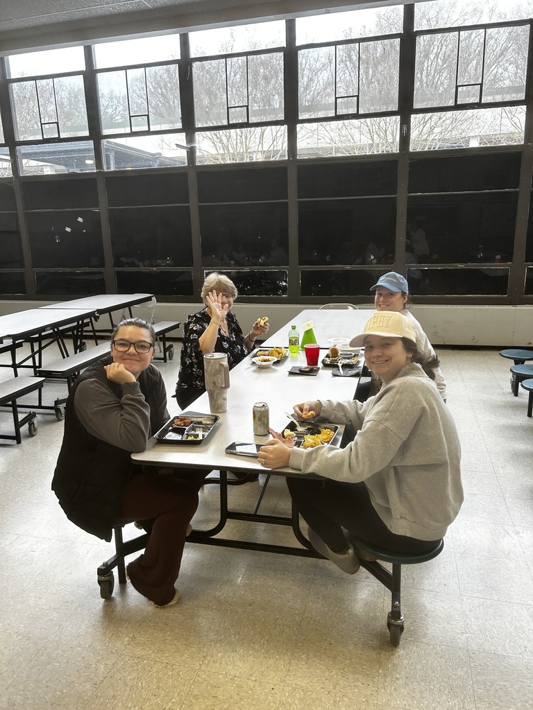 Four people sit around a white rectangular cafeteria table beneath large multi‑pane windows showing bare trees outside. From left: a dark‑haired woman with glasses (dark vest, grey shirt, maroon pants) smiles with her chin on her hand; across from her an older blonde woman in a patterned top waves and holds food; on the right two younger women in light baseball caps and sweatshirts smile while eating. Trays with partitioned food, red and green plastic cups, a soda can, a tumbler, and a smartphone sit on the table. Empty cafeteria tables and light tile flooring are visible in the background.