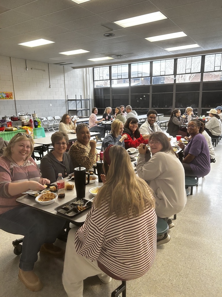 A busy communal meal in a large dining hall: about 20–30 adults sit at long tables with attached stools, eating and socializing under recessed ceiling lights and high multi‑pane windows that show trees outside. People in the foreground smile toward the camera; tables hold plates, trays, cups and bottles. Light cinderblock walls and a colorful wall decoration are visible, giving the scene a warm, social atmosphere.