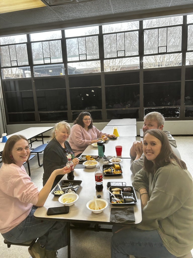 Five people sit along two long dining tables under large windows. On the left are three women (one in pink holding a bottle, an older blonde woman, and a dark‑haired woman eating from a tray). Facing the camera on the right are a smiling man in a light grey hoodie and a woman in an olive sweater. Multiple compartmented food trays, small bowls, plastic Coca‑Cola bottles, a red cup, and a teal thermos are on the tables. The mood is relaxed and friendly.
